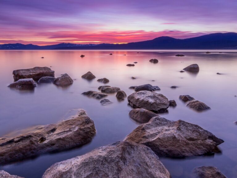 Calm relaxing scene of water reflecting purple sunset colours, with rocks sticking out. Mountains in background.