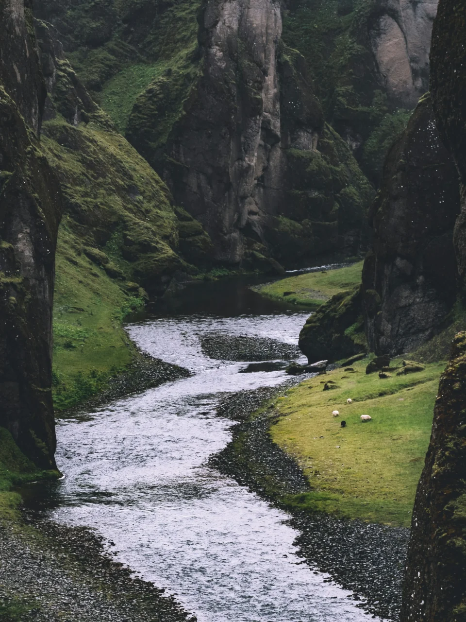 A flowing river flanked by grassy banks and mountainous cliffs.