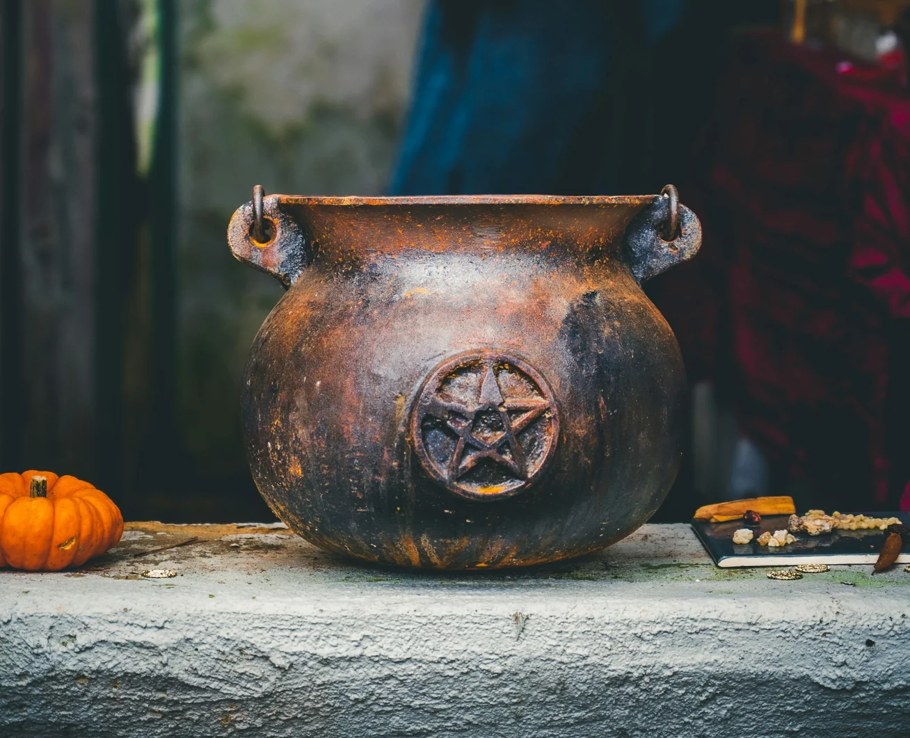 A weathered cauldron with a pentacle. Next to it, a small pumpkin.