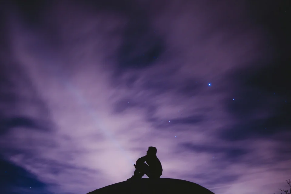 A silhouette of a person sitting, looking up at the purple sky.
