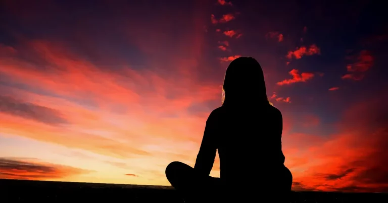 A silhouette of a person meditating as the sun sets. Warm reds and yellows in the sky.