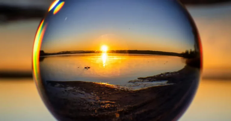 Sunset reflecting on calm water, seen through a glass sphere – symbolic of how we need to change perspective and allow self-reflection to guide us forward.