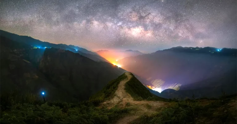 A mountain range at dusk, with small lights illuminating various paths. Ahead a path stretches up a peak, symbolic of our spiritual journeys.